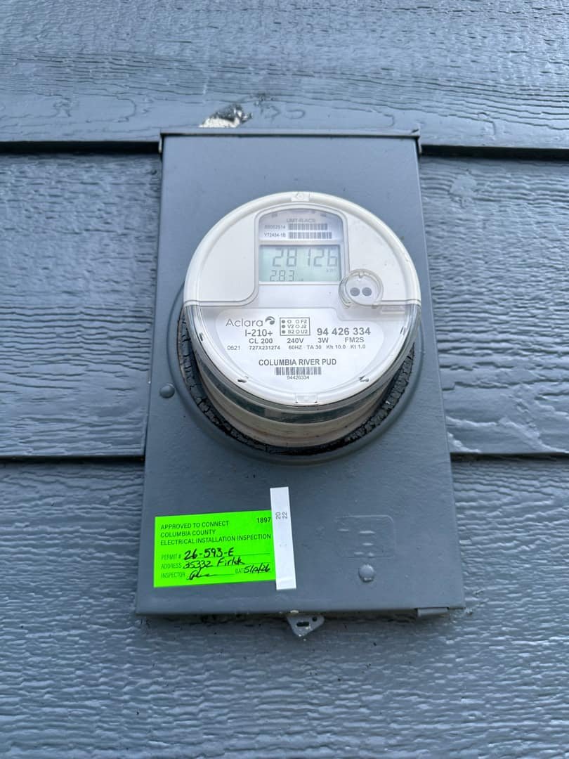 Digital electricity meter mounted on an exterior wall, with usage readings and inspection stickers visible.